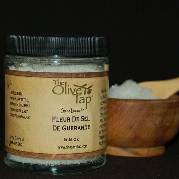 A jar of Fleur De Sel De Guerande Sea Salt from The Olive Tap with a wooden bowl and spoon full of sea salt in the background.
