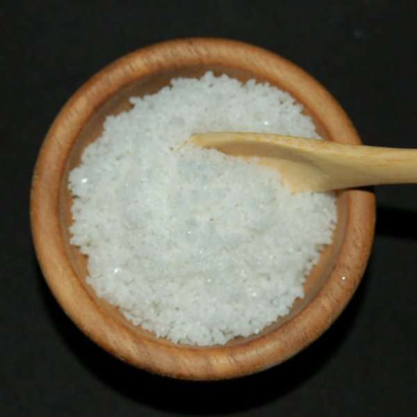 Fleur De Sel De Guerande Sea Salt from The Olive Tap in a wooden bowl with a wooden spoon against a black background.