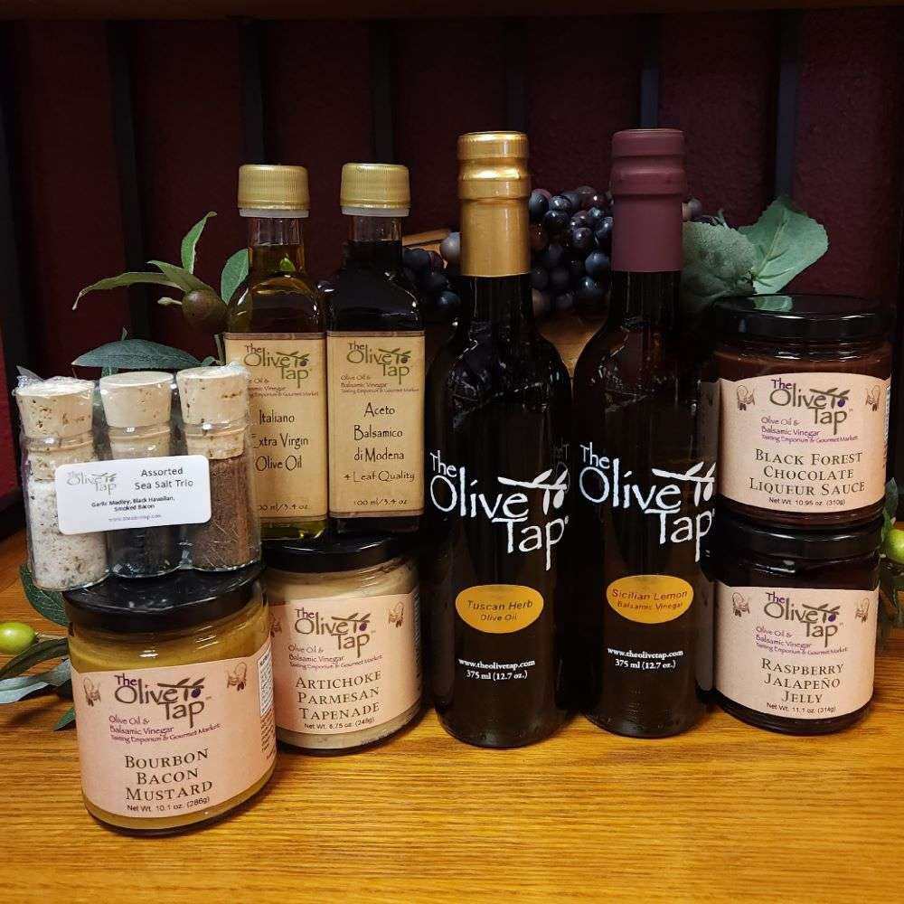 Foodie Frenzy Gift Box from The Olive Tap: One large bottle of olive oil, one large bottle of balsamic vinegar, one small bottle of olive oil, one small bottle of vinegar, liqueur sauce, jelly, mustard, tapenade, and a trio of assorted sea salts on a wooden surface with olive branches and purple grapes in the background.