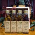 Four small bottles of Olive Tap olive oil and balsamic vinegar arranged in front of a gift box on a wooden surface. The bottles contain different flavored olive oils and balsamic vinegars: Blood Orange, Granny Smith, Sorrento Lemon, and Cranberry Pear.