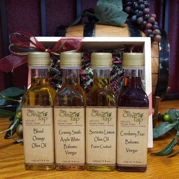 Four small bottles of Olive Tap olive oil and balsamic vinegar arranged in front of a gift box on a wooden surface. The bottles contain different flavored olive oils and balsamic vinegars: Blood Orange, Granny Smith, Sorrento Lemon, and Cranberry Pear.