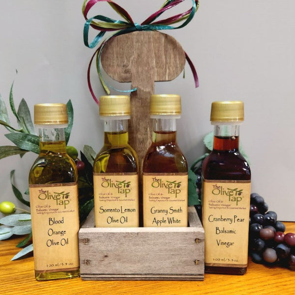 Four small glass bottles of olive oil and vinegar arranged in a wooden caddy crate. The bottles have gold caps and labels reading Blood Orange Olive Oil, Scented Lemon Olive Oil, Granny Smith Apple White Olive Oil, and Cranberry Pear Balsamic Vinegar.