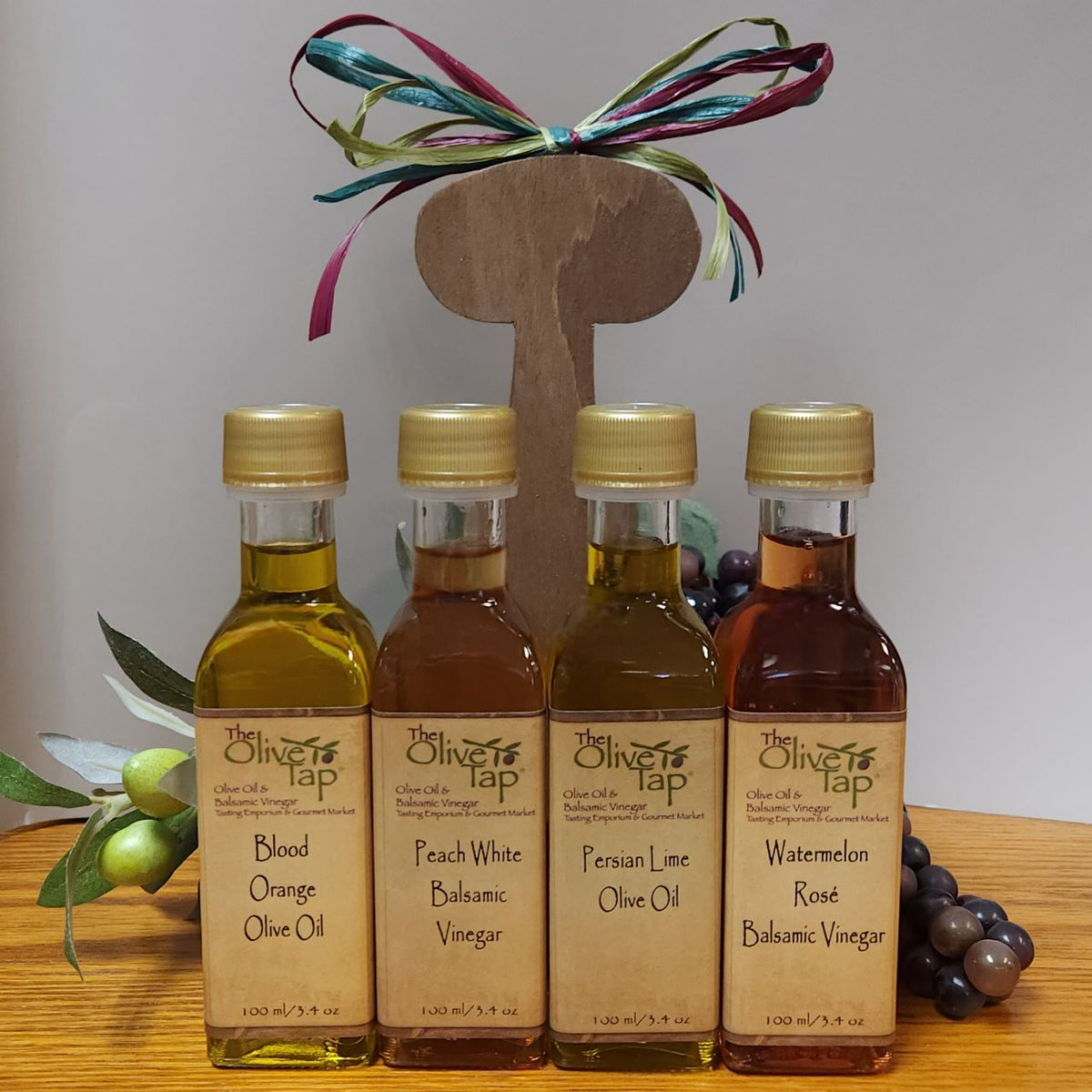 The Olive Tap's Fruity Sampler Caddy Summer Flavors: two olive oil bottles and two vinegar bottles in a wooden caddy with a bow on a wood surface and with olive branches and grapes in the background.