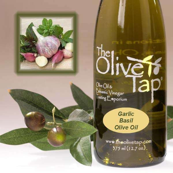A bottle of Garlic Basil Olive Oil from The Olive Tap next to olives and leaves with an inset showing bulbs of garlic and fresh basil leaves.
