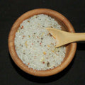Garlic Medley Sea Salt from The Olive Tap: White pebble-sized chunks of sea salt with green and orange flakes mixed in, in a wooden bowl with a wooden spoon inside of it, against a black background.