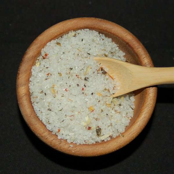 Garlic Medley Sea Salt from The Olive Tap: White pebble-sized chunks of sea salt with green and orange flakes mixed in, in a wooden bowl with a wooden spoon inside of it, against a black background.