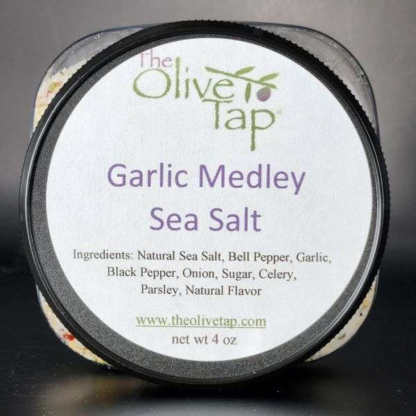 Garlic Medley Sea Salt In Jar from The Olive Tap with a white label that includes The Olive Tap logo with an olive branch, product name, and ingredients list, with the jar in front of a gray background.