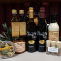 The Gourmet Sampler Gift Box from The Olive Tap: Olive oils, vinegars, seasoning, sauce, sea salts, and a tapenade in front of olive branches and purple grapes.