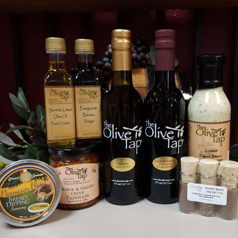 The Gourmet Sampler Gift Box from The Olive Tap: Olive oils, vinegars, seasoning, sauce, sea salts, and a tapenade in front of olive branches and purple grapes.