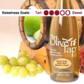 A bottle of Granny Smith Apple White Balsamic Vinegar from The Olive Tap surrounded by grapes in front of a wooden barrel with granny smith apples inside, and a sweetness scale indicating a three on a light background.