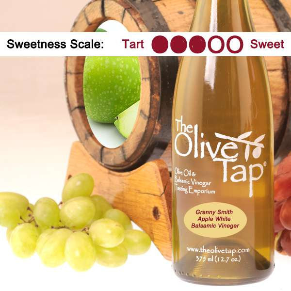A bottle of Granny Smith Apple White Balsamic Vinegar from The Olive Tap surrounded by grapes in front of a wooden barrel with granny smith apples inside, and a sweetness scale indicating a three on a light background.