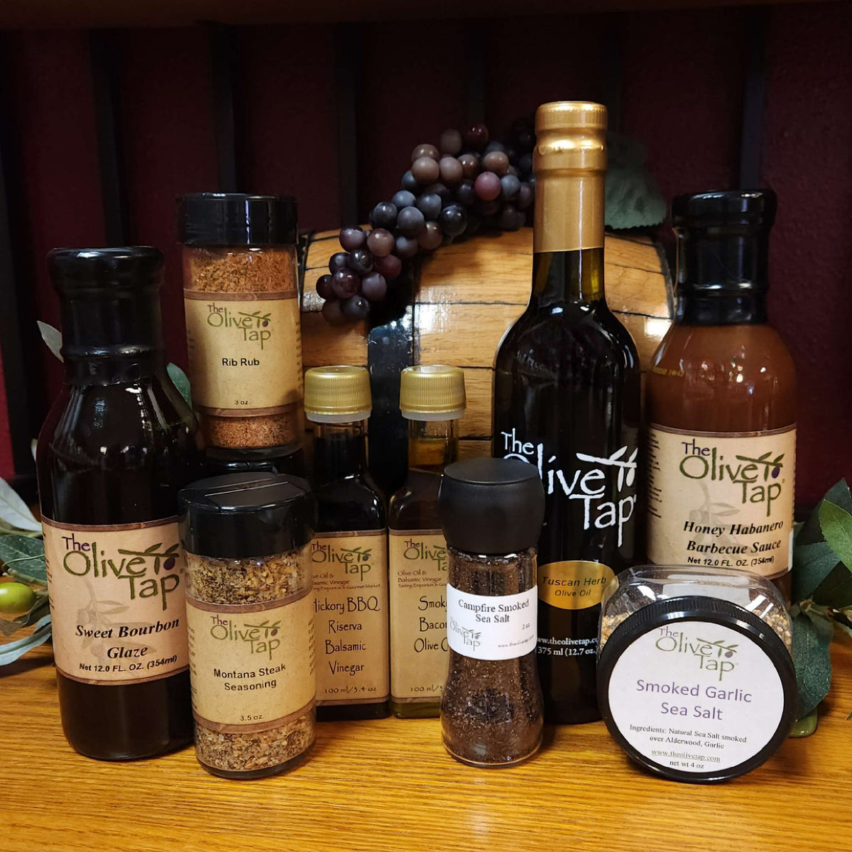 Grill Master Gift Box from The Olive Tap: Bottles of olive oil, a sauce, a glaze, and a rub, seasoning, and sea salts on a wooden surface in front of a wooden barrel with grapes and surrounded by olives and leaves.