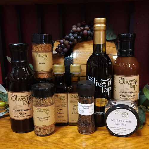 Grill Master Gift Box from The Olive Tap: Bottles of olive oil, a sauce, a glaze, and a rub, seasoning, and sea salts on a wooden surface in front of a wooden barrel with grapes and surrounded by olives and leaves.
