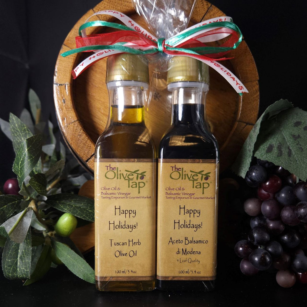 Happy Holidays Oil and Vinegar Pairing from The Olive Tap: A bottle of olive oil and a bottle of vinegar with a holiday bow on top of them with a decorative wooden barrel, purple grapes, and an olive branch in the background.