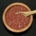 Hawaiian Red Sea Salt from The Olive Tap: Pebble-sized red pieces of sea salt  in a wooden bowl with a wooden spoon against a black background.
