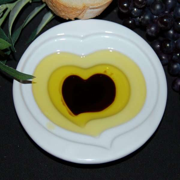 Pinzimonio Olive Oil Dipping Dish - Heart Design from The Olive Tap with olive oil and vinegar inside and a slice of bread with grapes and olive leaves in the background.