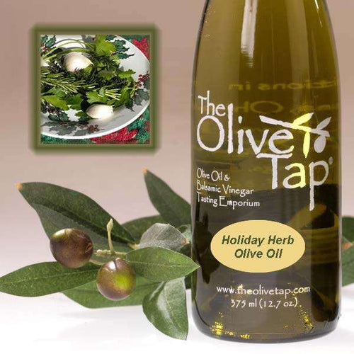 A bottle of Holiday Herb Olive Oil from The Olive Tap next to olives and leaves with an inset showing fresh herbs on a light background.