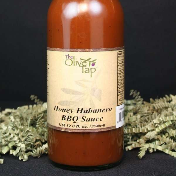 A bottle of Honey Habanero BBQ Sauce from The Olive Tap with green crinkle paper against a black background.