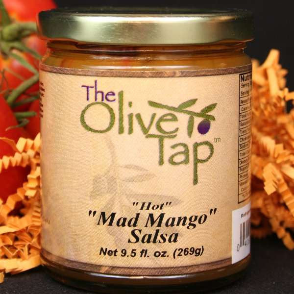 A jar of Mad Mango Salsa from The Olive Tap, surrounded by orange decorative crinkle paper and fresh tomatoes on vines in the background, all on a black surface.
