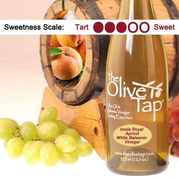 A bottle of Imola Royal Apricot White Balsamic Vinegar from The Olive Tap surrounded by grapes in front of a wooden barrel with apricots inside, and a sweetness scale indicating a three on a light background.