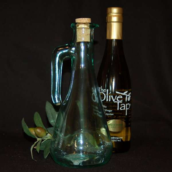 Italian Glass Cruet (17 oz.) from The Olive Tap: A bottle of olive oil from The Olive Tap next to a decorative empty olive oil bottle with a cork in front of olives and leaves on a dark background.