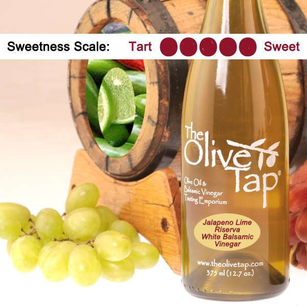 A bottle of Jalapeno Lime Riserva White Balsamic Vinegar surrounded by grapes in front of a wooden barrel with limes and jalapenos inside, and a sweetness scale indicating a five on a light background.