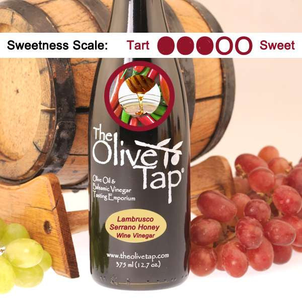 A bottle of Lambrusco Serrano Pepper Honey Vinegar 375ml bottle from The Olive Tap with a picture of drizzled honey and serrano peppers, surrounded by grapes in front of a wooden barrel, and a sweetness scale indicating a three on a light background.