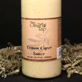 A bottle of Lemon Caper Sauce from The Olive Tap surrounded by green decorative crinkle paper against a black background and surface.