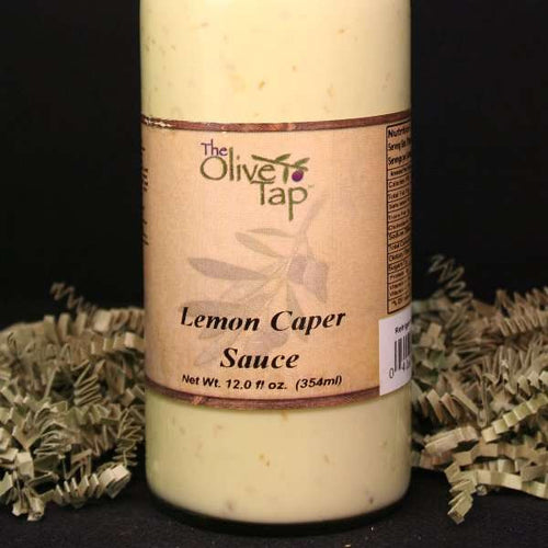 A bottle of Lemon Caper Sauce from The Olive Tap surrounded by green decorative crinkle paper against a black background and surface.