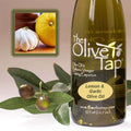 A bottle of Lemon and Garlic Olive Oil from The Olive Tap next to olives and leaves with an inset showing a garlic and a lemon on a light background.