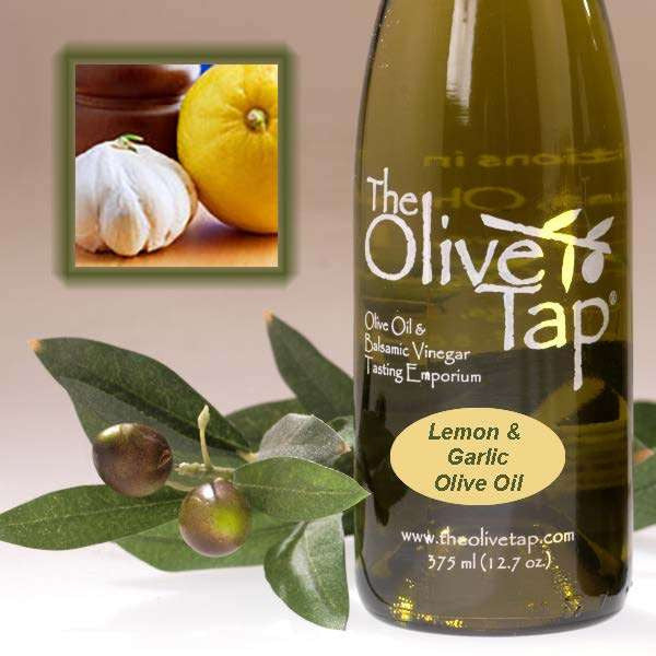 A bottle of Lemon and Garlic Olive Oil from The Olive Tap next to olives and leaves with an inset showing a garlic and a lemon on a light background.