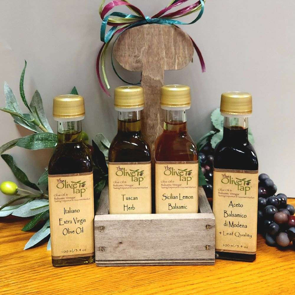 Little Italy in Caddy with Standard Trim from The Olive Tap: A caddy topped with a bow with olive oil and balsamic vinegar bottles inside and two more bottles, one on each side, on the outside of the caddy, all on a wooden surface in front of grapes and olives with leaves.