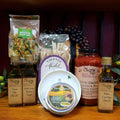 Mangia Italian Dinner Gift Box from The Olive Tap: A tin of seasoning, two bottles of olive oil, one bottle of vinegar, a jar of pasta sauce, bread dipping plate, package of pasta, and wedding soup mix on a wooden surface with olive branches and purple grapes in the background.