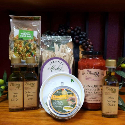 Mangia Italian Dinner Gift Box from The Olive Tap: A tin of seasoning, two bottles of olive oil, one bottle of vinegar, a jar of pasta sauce, bread dipping plate, package of pasta, and wedding soup mix on a wooden surface with olive branches and purple grapes in the background.