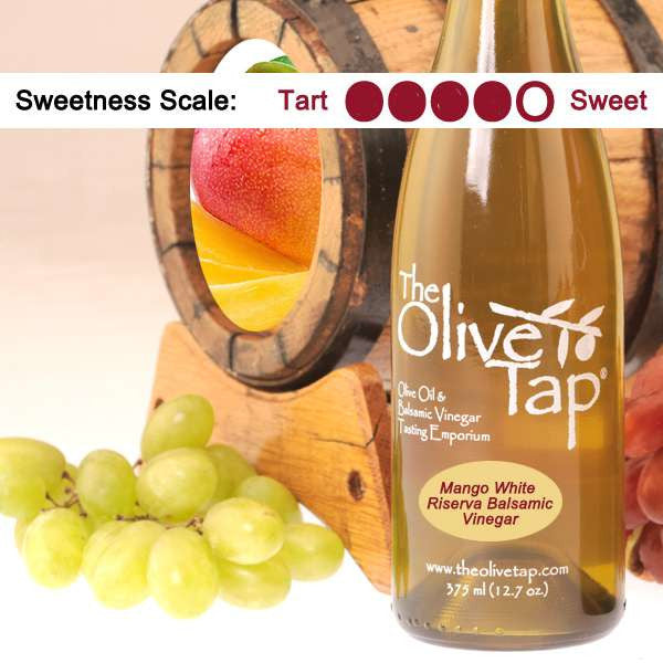A bottle of Mango White Riserva Balsamic Vinegar from The Olive Tap surrounded by grapes in front of a wooden barrel with a picture of a mango inside, and a sweetness scale indicating a four on a light background.