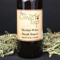 A bottle of Merlot Wine Steak Sauce from The Olive Tap, surrounded by decorative green crinkle paper against a black background and surface.