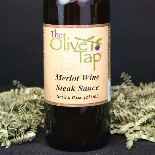 A bottle of Merlot Wine Steak Sauce from The Olive Tap, surrounded by decorative green crinkle paper against a black background and surface.
