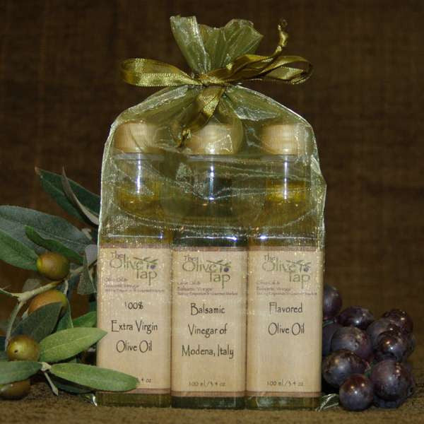 Tap's Trio Oil and Vinegar Sampler Sets in Sachet Bag from The Olive Tap: Two bottles of olive oil and a bottle of vinegar in an organza bag with a bow beside grapes, olives, and leaves on a brown backdrop.
