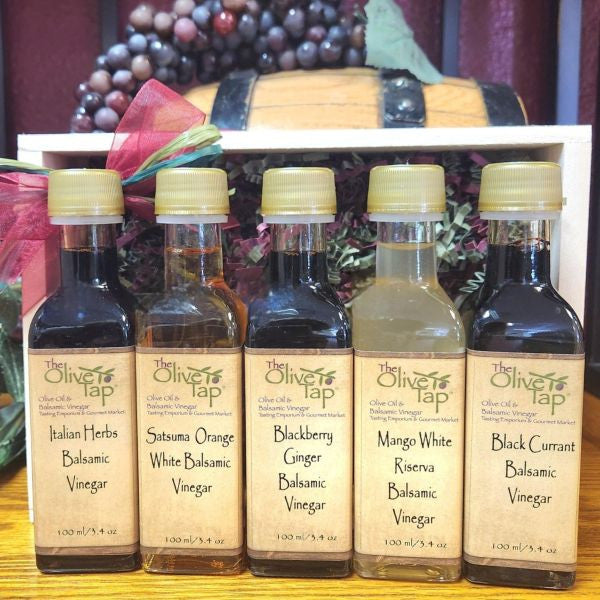 The Olive Tap Vinegar Sampler from The Olive Tap: Five bottles of vinegar lined up in a wooden box with a bow on a wooden surface in front of a decorative wooden barrel with grapes on top.