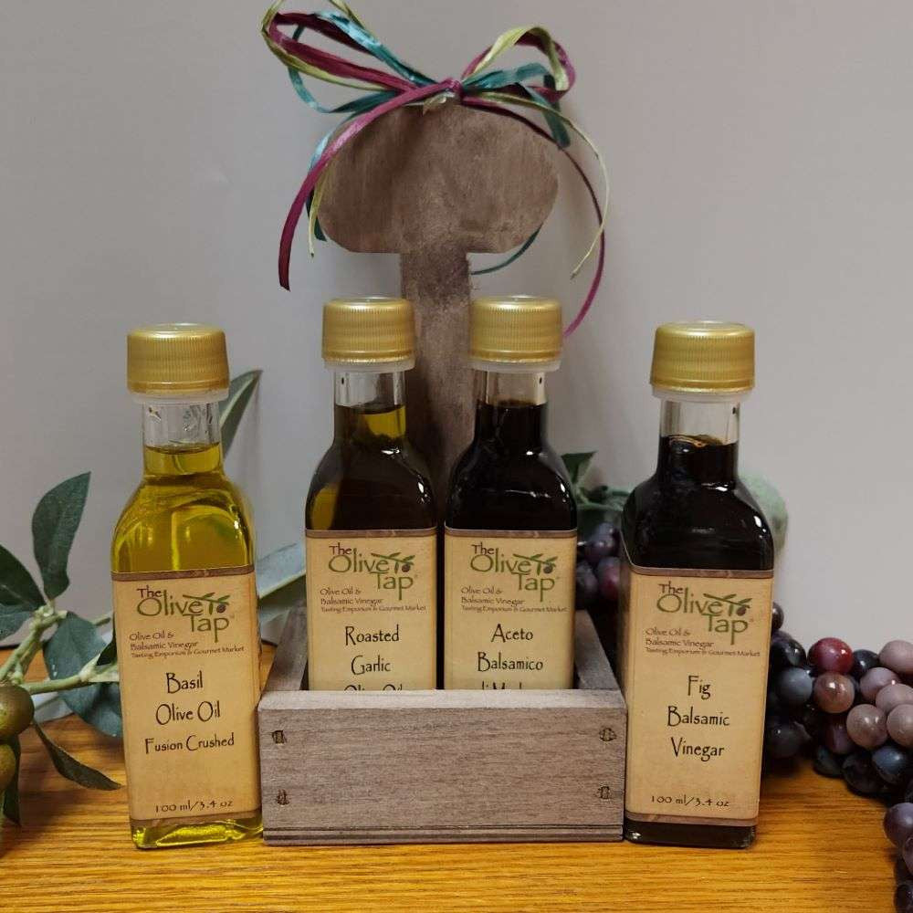 Originale Sampler in Caddy from The Olive Tap: A caddy with a bow on top with an olive oil and vinegar bottle inside with two more bottles, one on each side, on the outside, and the caddy is surrounded by olives, leaves, and grapes with a light background.