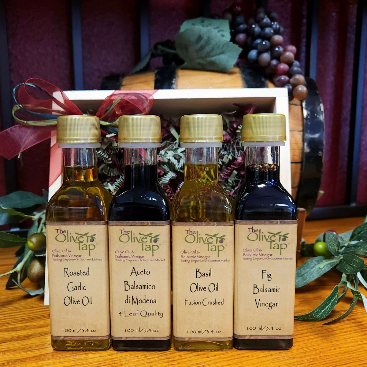 Originale Sampler by The Olive Tap: Two olive oil and two vinegar bottles in front of a wooden crate with a decorative bow, olives and branches, purple grapes, and a wooden barrel.