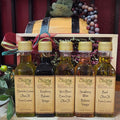 The Olive Tap Sampler Gift Set from The Olive Tap: Five Olive Tap bottles of olive oils and vinegars in front of a wooden crate with a bow and a wooden barrel with grapes.
