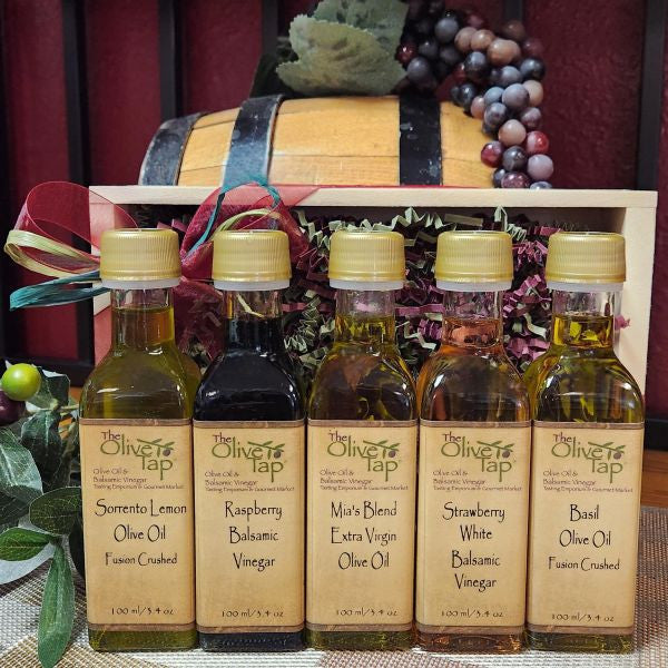 The Olive Tap Sampler Gift Set from The Olive Tap: Five Olive Tap bottles of olive oils and vinegars in front of a wooden crate with a bow and a wooden barrel with grapes.