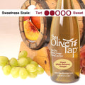 A bottle of The Olive Tap Peach White Balsamic Vinegar with peaches and grapes in front of a wooden barrel indicating a sweetness scale of two.
