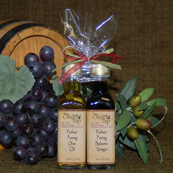 Perfect Pairing Sampler Set with Standard Trim from The Olive Tap: A bottle of olive oil and a bottle of vinegar with a bow on top, surrounded by grapes, olives, leaves, and a wooden barrel on a brown background.