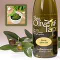 A bottle of Pesto Olive Oil from The Olive Tap next to olives and leaves with an inset showing a ramekin with pesto, a wooden spoon, fresh basil, and garlic on a light background.