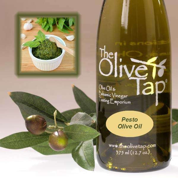 A bottle of Pesto Olive Oil from The Olive Tap next to olives and leaves with an inset showing a ramekin with pesto, a wooden spoon, fresh basil, and garlic on a light background.