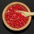 Pink Peppercorns from The Olive Tap in a wooden bowl with a wooden spoon in it against a black background.