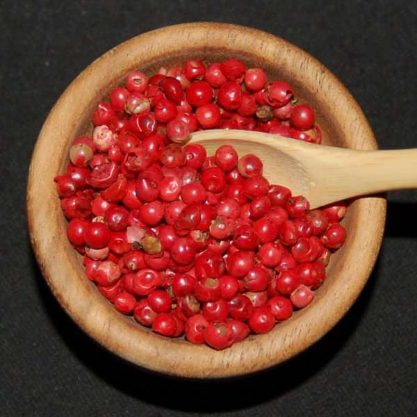Pink Peppercorns from The Olive Tap in a wooden bowl with a wooden spoon in it against a black background.