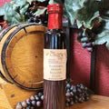 A bottle of Raspberry Merlot Balsamic Glaze from The Olive Tap in front of a decorative wooden barrel, purple grapes and their leaves, and a red wall in the background.